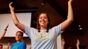 A young woman with brown skin smiles and raises her arms in an upward arc.