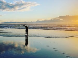A figure of a person on a beach with water reflecting the sky, reaching upwards in freedom.