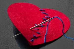 A red fabric heart stitched together by blue thread and a needle.